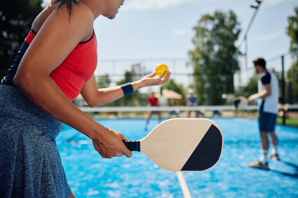 A girl is playing pickleball in a red shirt and blue shorts while holding a yellow ball and pickleball paddle