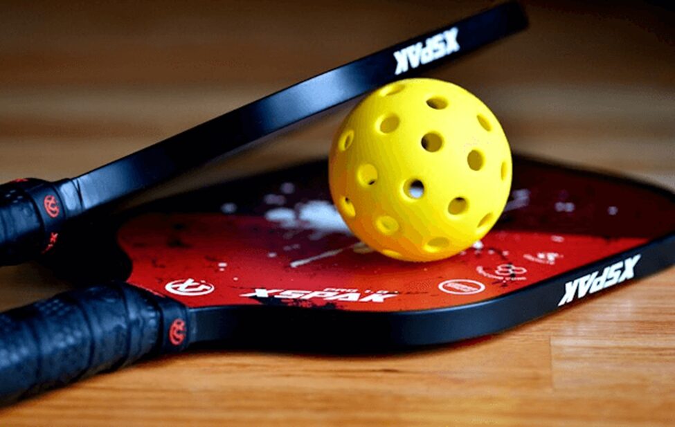 It shows a pickleball and paddle made of yellow color, both of which are used in playing a pickleball sports